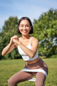 Asian fitness girl doing squats in park, using resistance band, stretching yoga rope for workout training on fresh air.